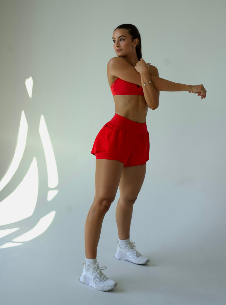 The coah is posing in a studio with red workout outfit while streching her arm..
